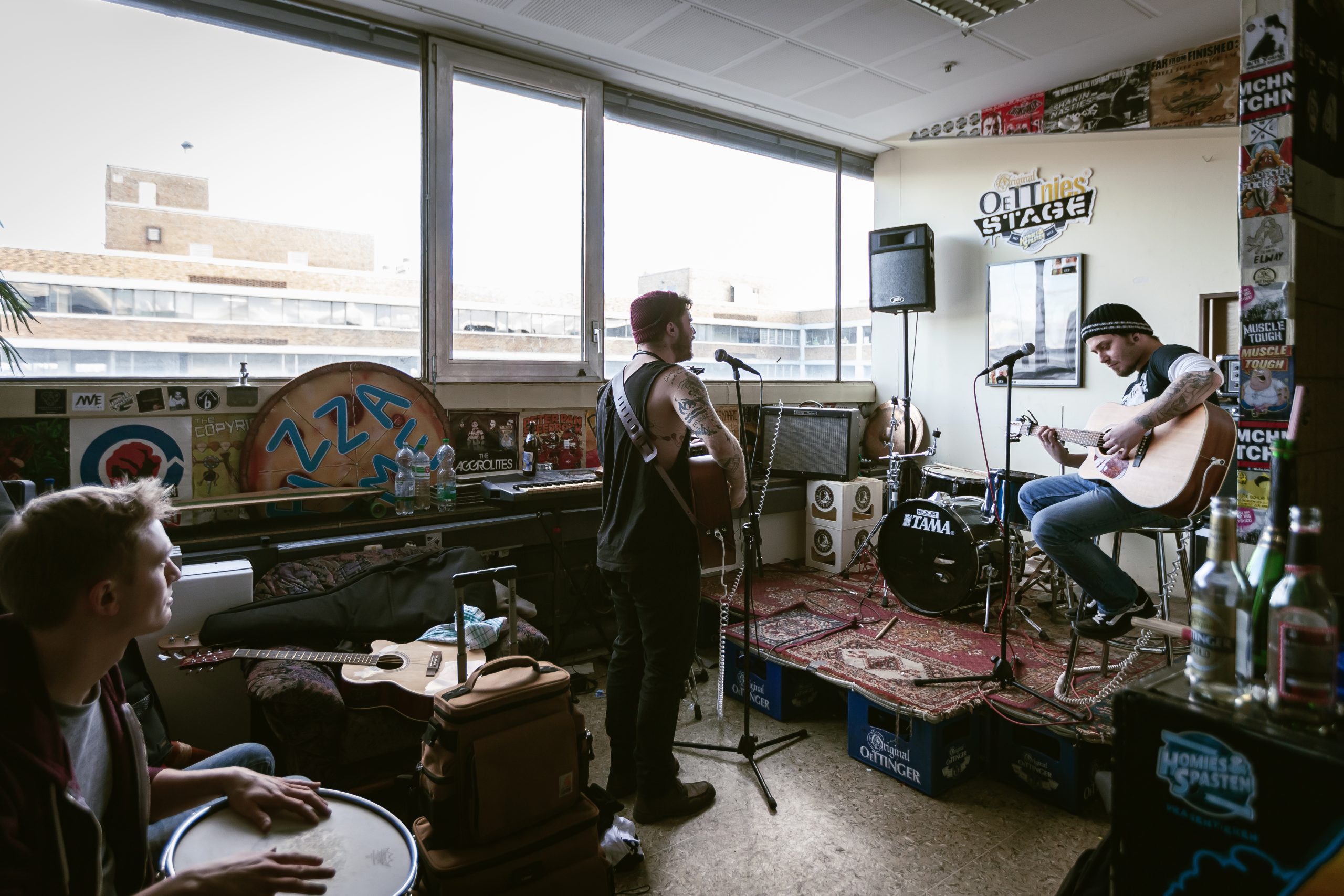 Zwei Musiker proben in einem Raum, eine Person sitzt mit Gitarre auf einem Stuhl. Im Hintergrund sind große Fenster, die den Blick auf einen Teil des Bestandsgebäudes The Q eröffnen.