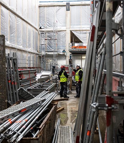 Bild einer Baustelle mit zwei Personen in gelben Schutzwesten. Auf dem Boden lagern Stahlelemente als Baumaterial.